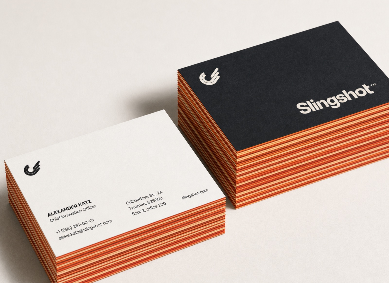 Two stacks of Slingshot business cards: a Carbon face with the Warm Stone mark and wordmark, a Warm Stone face with the Carbon mark, both with layered rust and amber painted edges